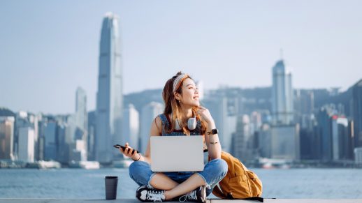 Cheerful young Asian woman sitting cross-legged by the promenade, against urban city skyline. She is wearing headphones around neck, using smartphone and working on laptop, with a coffee cup by her side. Looking away with smile. Lifestyle and technology