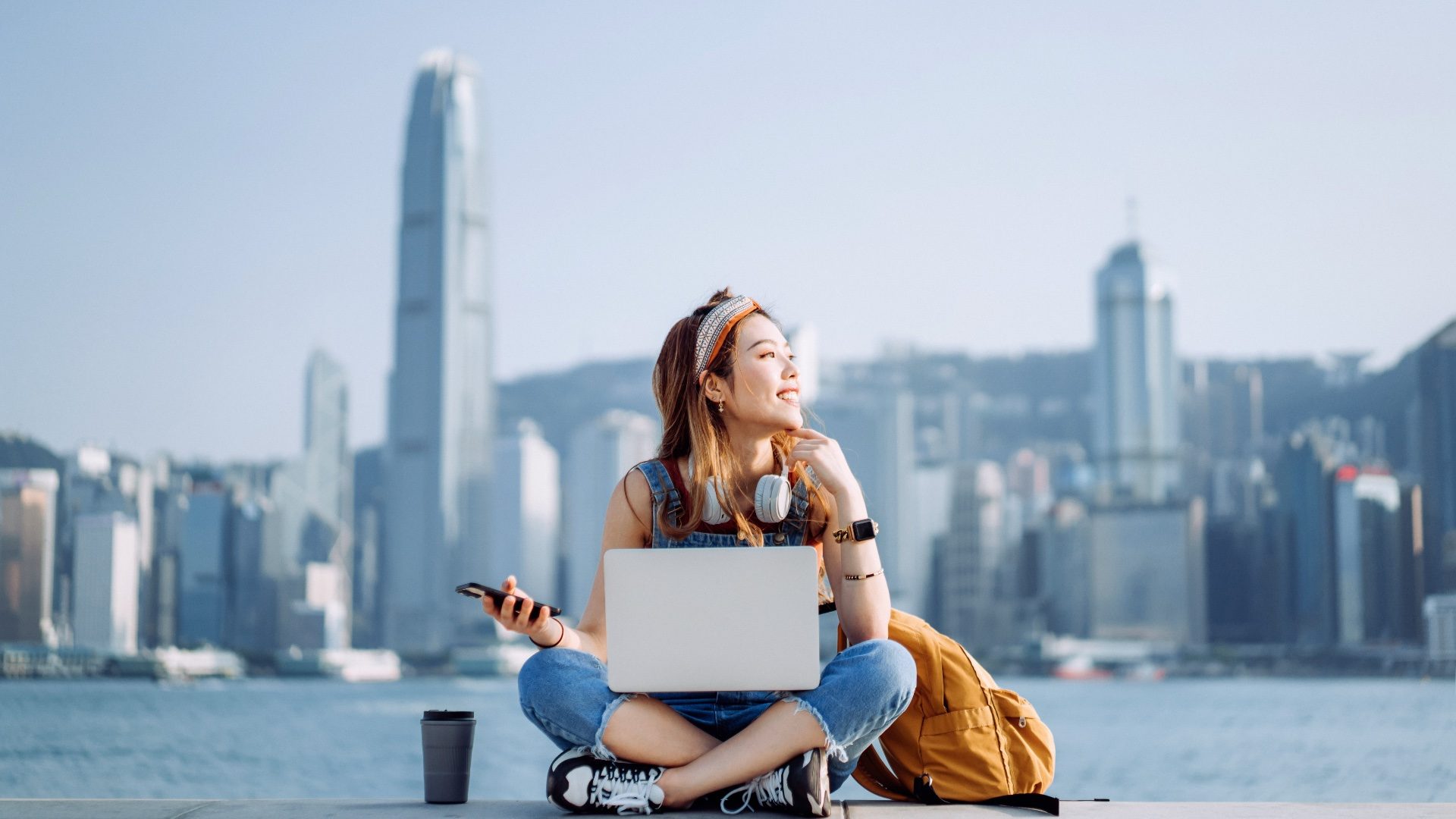 Cheerful young Asian woman sitting cross-legged by the promenade, against urban city skyline. She is wearing headphones around neck, using smartphone and working on laptop, with a coffee cup by her side. Looking away with smile. Lifestyle and technology
