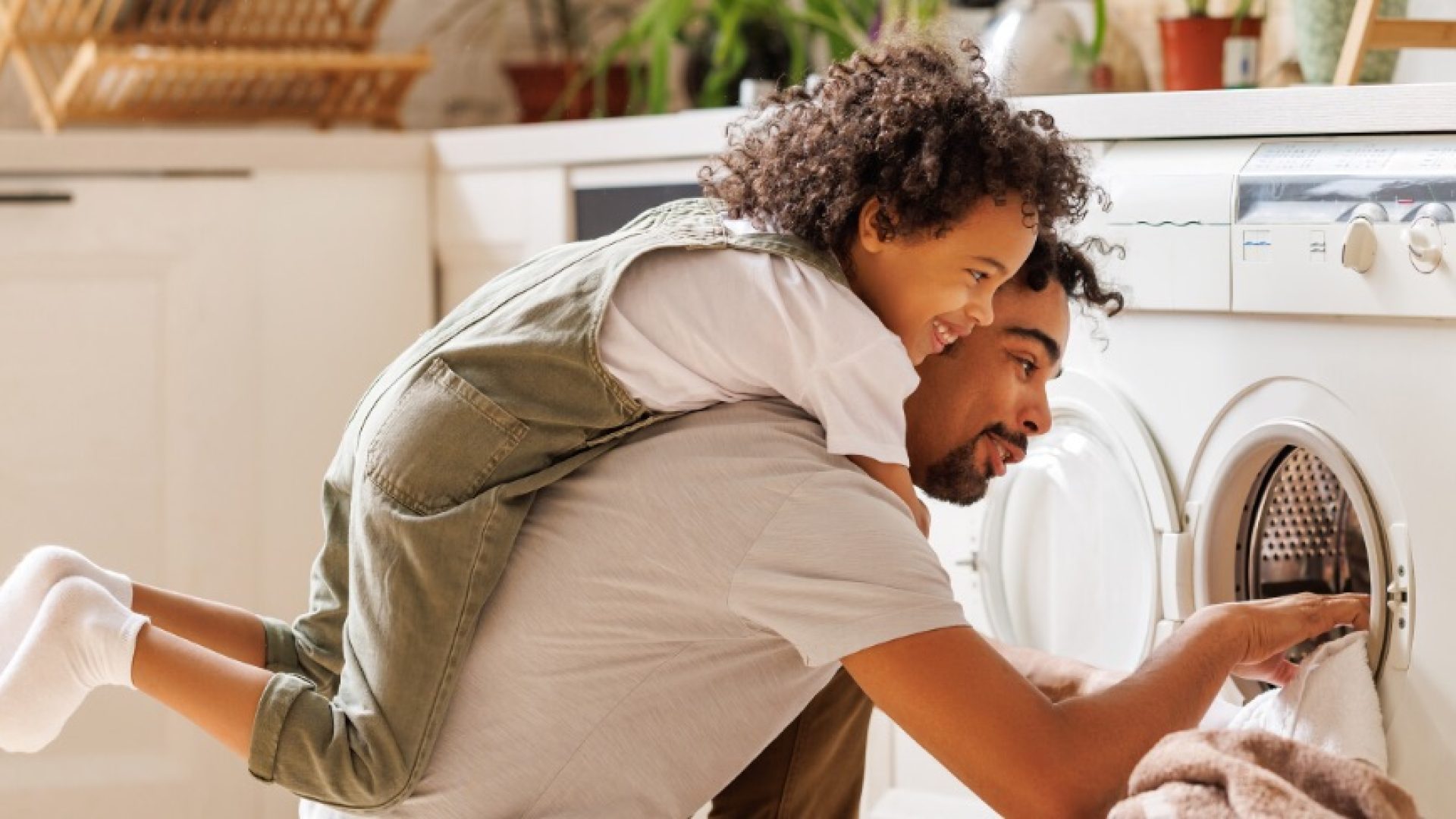 Side view of black child in casual clothes with curly hair smiling and embracing dad loading washing machine during household routine in morning at home; Shutterstock ID 2155189331; purchase_order: 1106262587; job: ; client: ; other: