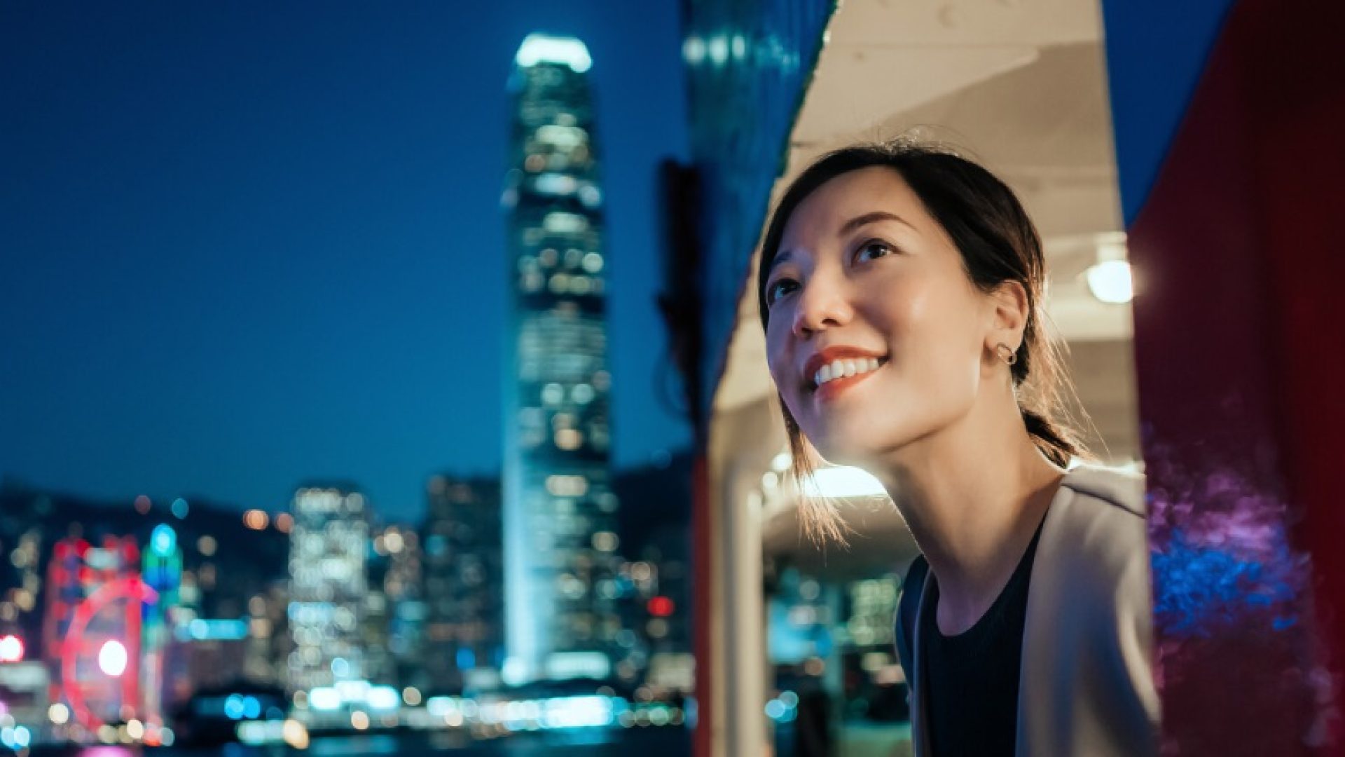 Beautiful smiling young Asian woman looking out through the window, enjoying spectacular illuminated night view of the city while travelling by ferry