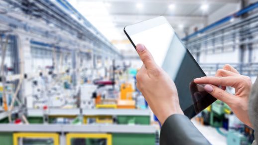 Horizontal color image of businesswoman - unrecognizable person - working with digital tablet in large futuristic factory. Woman standing on top of a balcony, holding touchpad and checking inventory of a manufacturing company on touchscreen tablet. Focus on businesswoman's hands holding black tablet, futuristic machines in background.