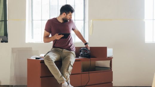 A young Caucasian man holding a digital tablet while sitting on a chest of drawers during a break from restoring furniture