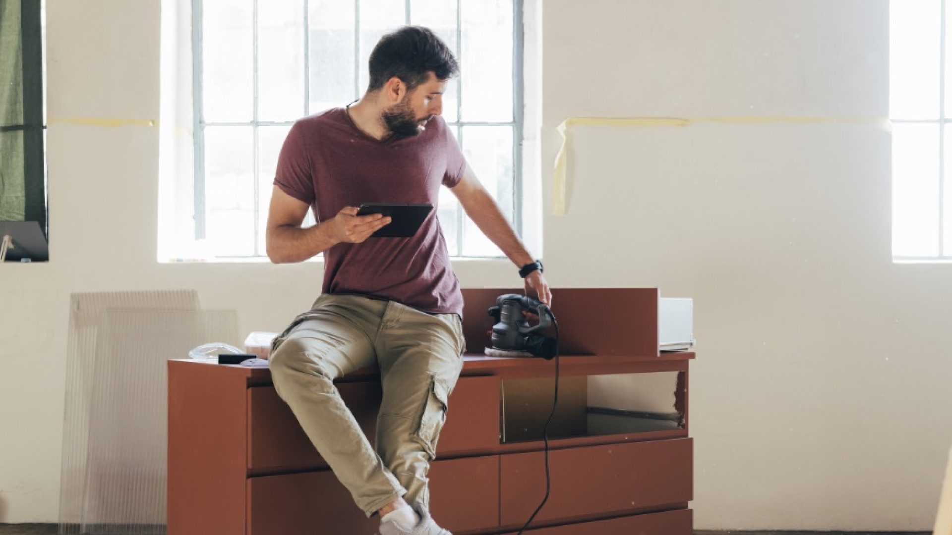 A young Caucasian man holding a digital tablet while sitting on a chest of drawers during a break from restoring furniture