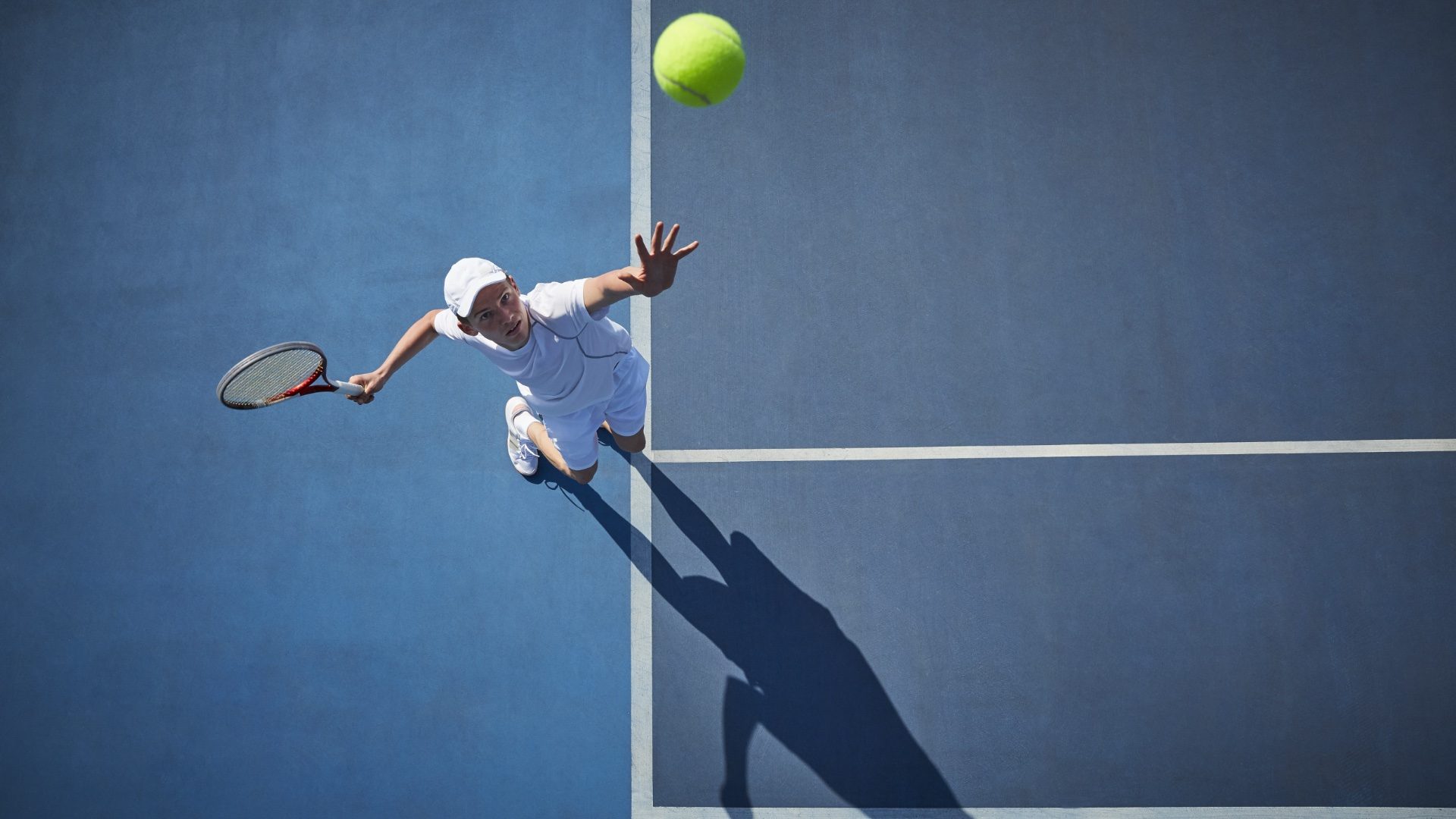 Overhead view of young male tennis player playing tennis, serving the ball on sunny blue tennis court